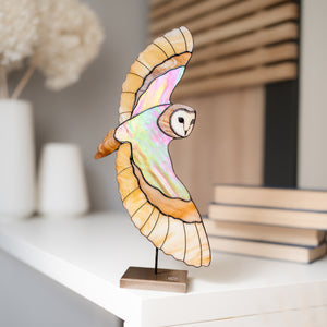A decorative owl sculpture with spread wings, shown on a small wooden base in a softly styled indoor setting. The owl has iridescent pastel wing panels outlined in dark lines, with a white and brown face, and stands on a light wood base.