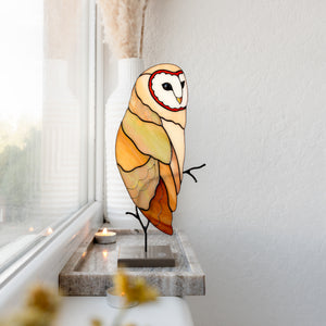 A stained-glass-style owl wall light in warm cream, yellow, orange, and brown tones with black outlines, displayed on a small stand on a windowsill in a real interior setting.