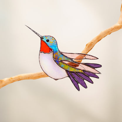 A stained glass cutout of a hummingbird perched on a branch, featuring iridescent blue and green wings, a bright red chest, and purple tail feathers, set against a soft beige background.