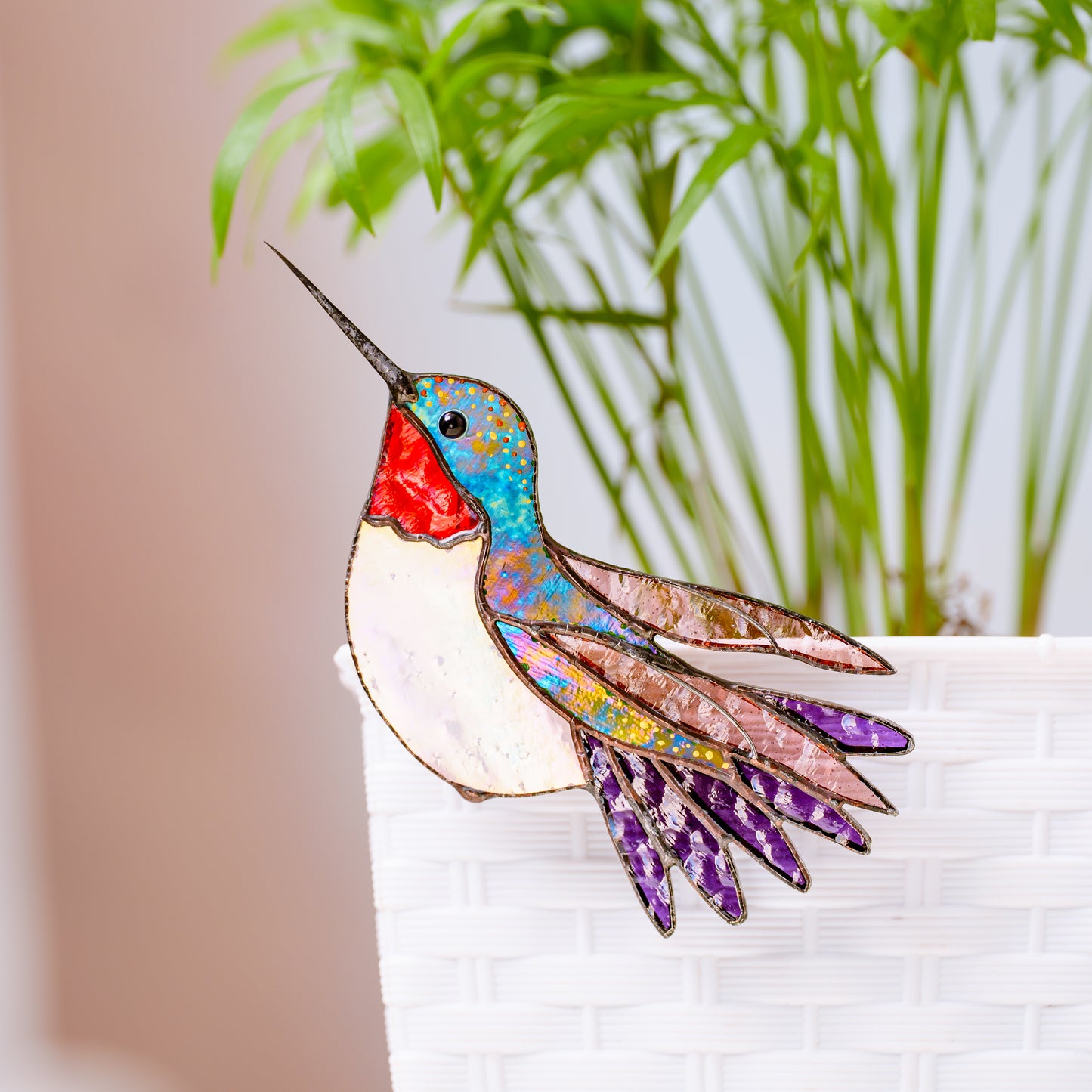 A stained glass hummingbird ornament with a blue, red, white, and purple body, perched on the edge of a white brick planter against a soft-focus background of green plants.