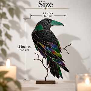 A stained-glass-style raven sculpture on a small branch stand, shown with overlaid size measurements reading 7 inches wide by 12 inches tall. The raven has a black body with green, blue, purple, and gold iridescent sections, photographed indoors against a light wall with candles blurred in the foreground.