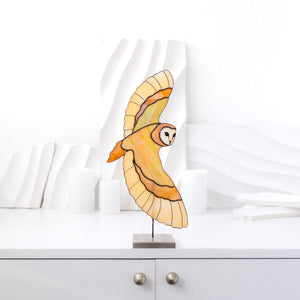 A stained glass owl sculpture with outstretched wings, mounted on a small black stand on a white cabinet. The background features a white wavy decorative wall and soft-focus white objects.